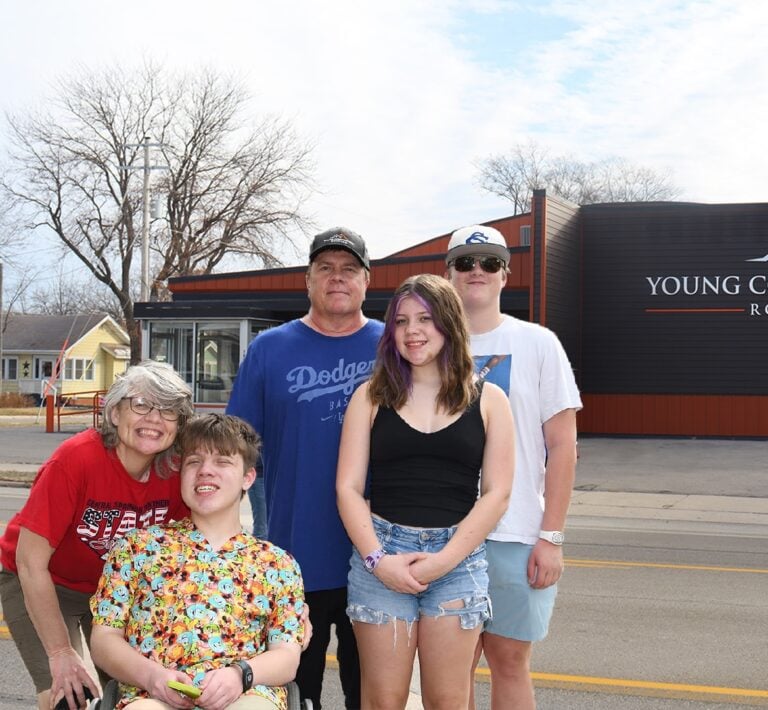 A group of five people, including one individual in a wheelchair wearing a floral shirt, pose and smile on the street in front of the Young County Arena—constructed by Young Construction. The remaining individuals stand behind in casual summer attire. Trees and houses are visible in the background.