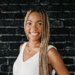 Smiling woman with long braids, white sleeveless top, and gold necklace stands before black brick wall on Hook Agency website.