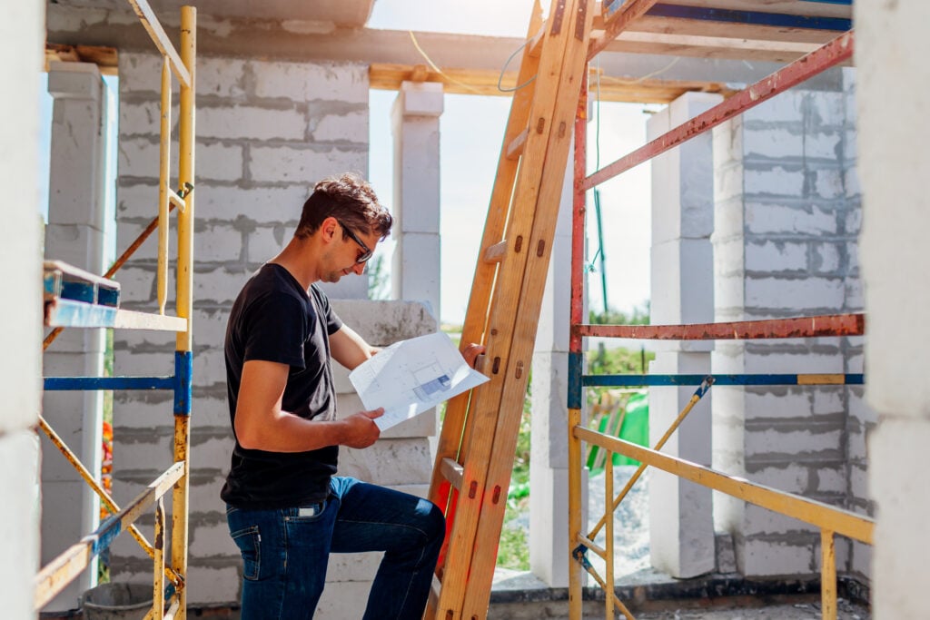 Leader in black shirt reviews blueprints atop ladder at active construction site, surrounded by scaffolding and brick walls.