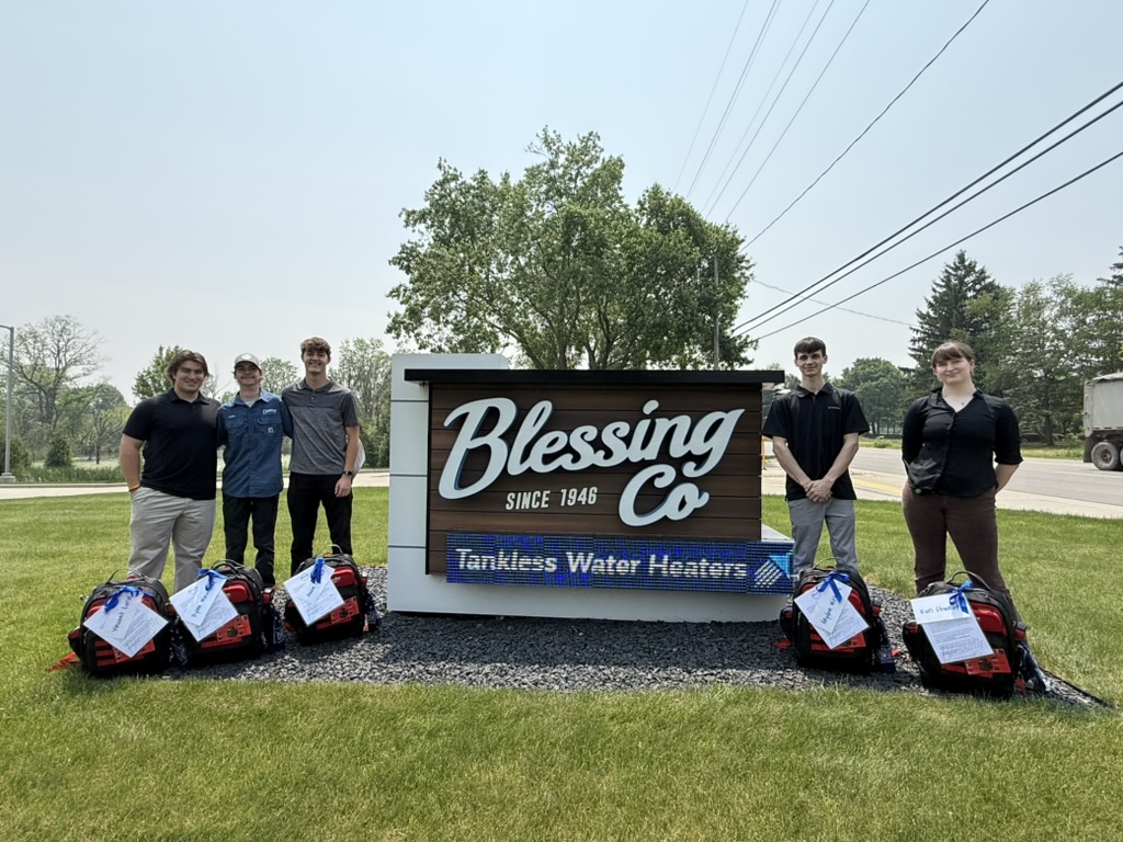 Six team members stand by a “Blessing Co Tankless Water Heaters” sign, wearing branded backpacks, with company vehicles behind.