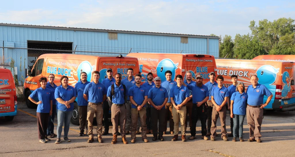 Blue Duck Plumbing team in blue shirts stands before branded orange vans outside an industrial building on a sunny day.