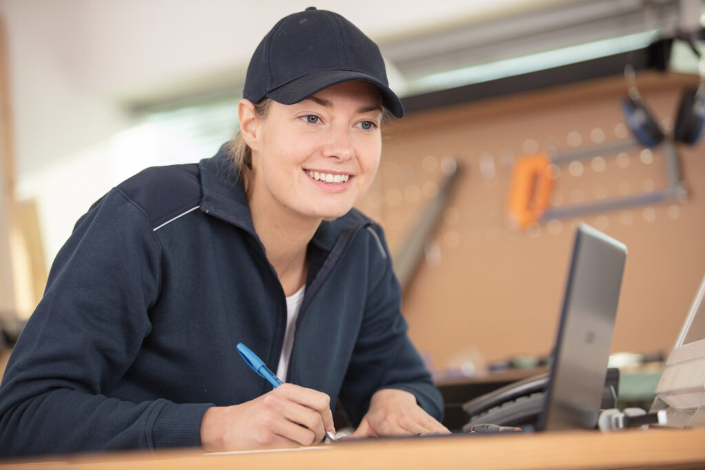 Professional in a navy cap and jacket smiles while planning home service website improvements at a desk with laptop and notepad.