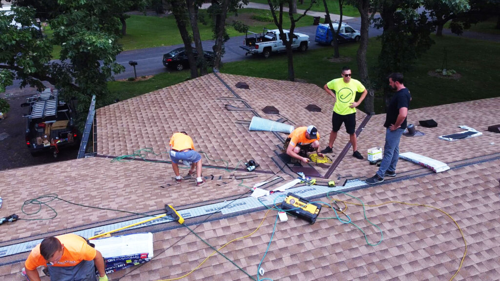 Roofing crew installs shingles and equipment on large brown roof, showcasing leadership. Tools, trees, vehicles in background.
