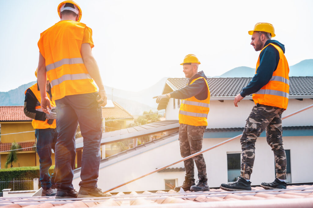 Four construction workers in safety gear discuss leadership on a rooftop, gesturing toward homes and mountains in bright sunlight.