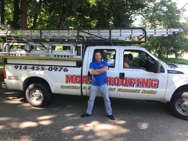 A man stands with arms crossed before a "MCAS ROOFING" truck, showcasing industry expertise. Another person sits inside.