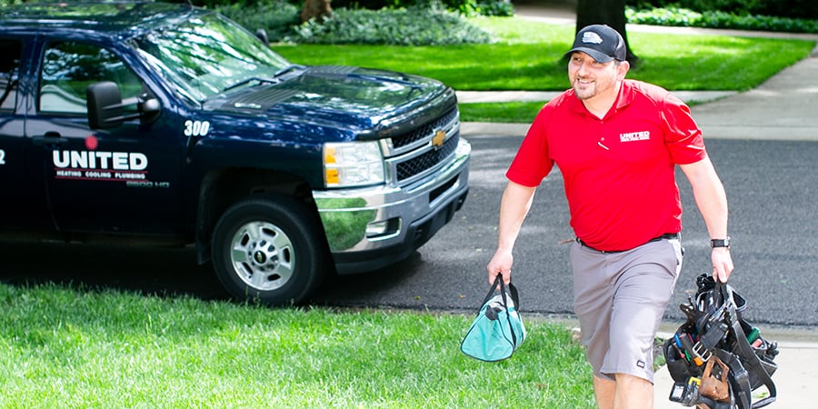 A professional United technician in red uniform carries tool bags on a lawn, with branded service truck parked nearby on the street.