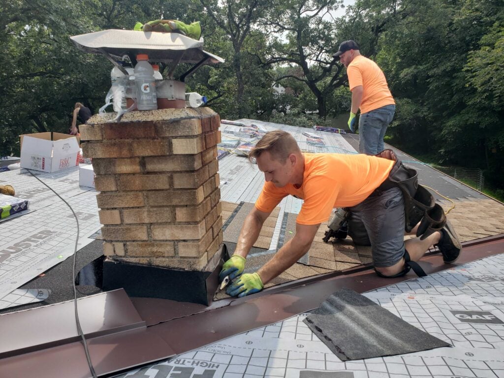 Two skilled roofers in orange shirts, one fitting chimney flashing and one with tools, work efficiently on a rooftop. Trees behind.