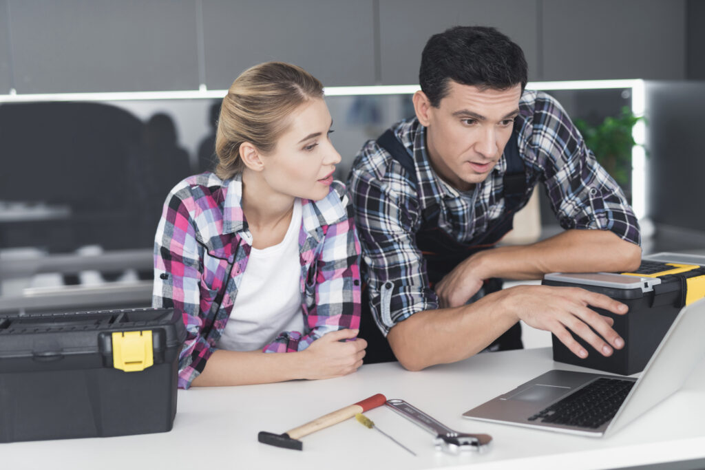 A woman and man in plaid shirts review their marketing budget for home repair services on a laptop at a kitchen counter with tools.
