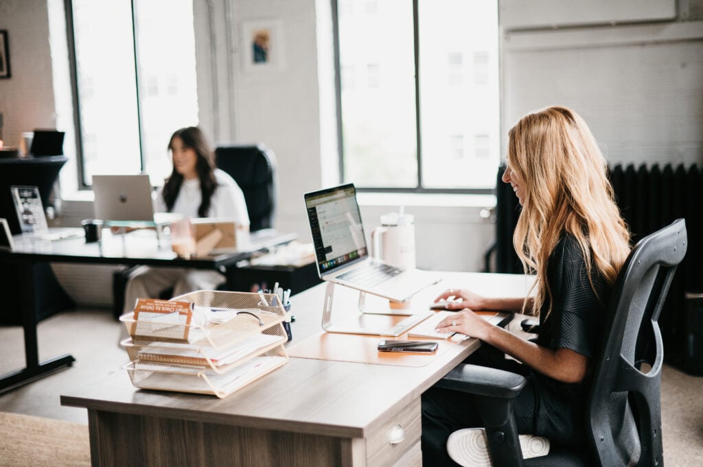 In a bright, modern office, two women work at separate desks—one typing, one researching top SEO companies for 2026.