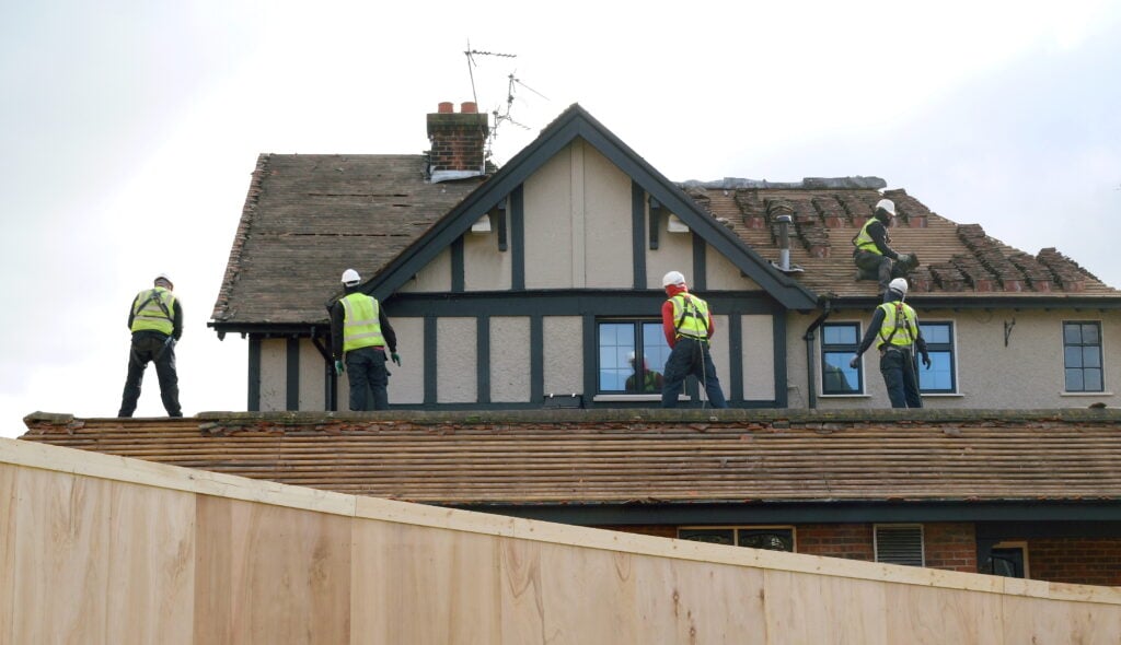 Five roofers in safety gear install shingles on a two-story home, showcasing professional roofing work and equipment in use.