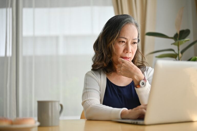 A woman reviews her laptop, considering Hook Agency’s free website audit. Sunlit workspace with mug, plate, and plant nearby.