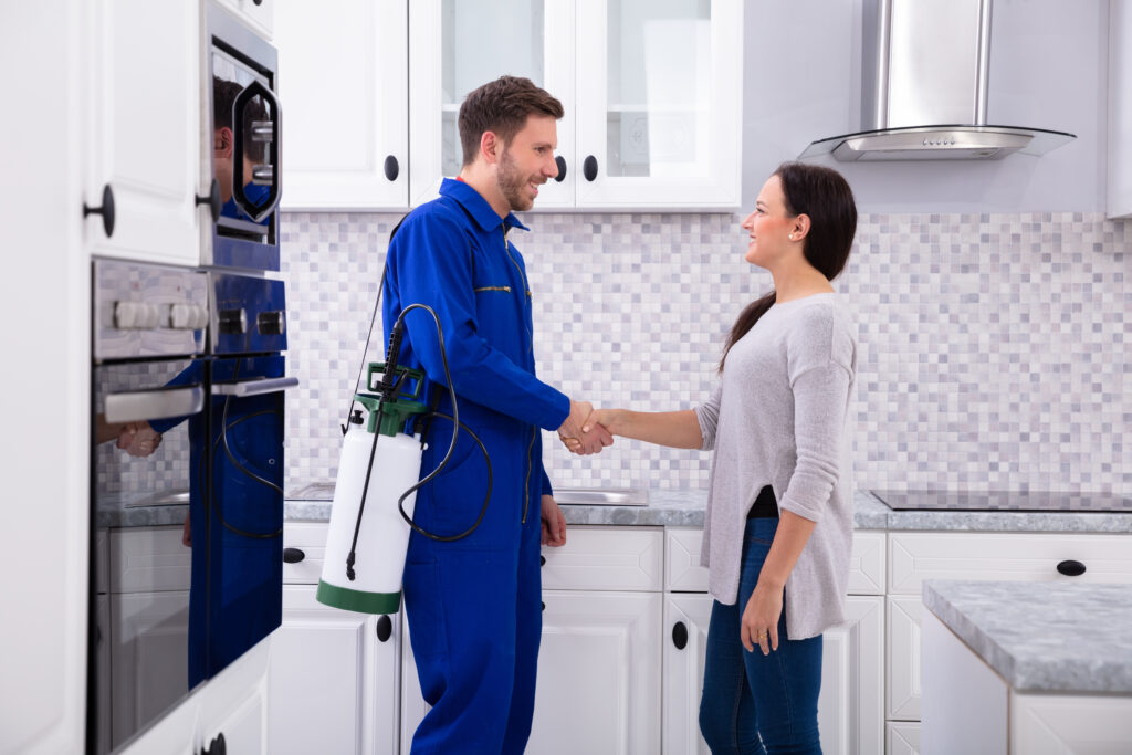 A pest control worker in blue uniform shakes hands with a smiling client in her kitchen, demonstrating professional, trusted service.