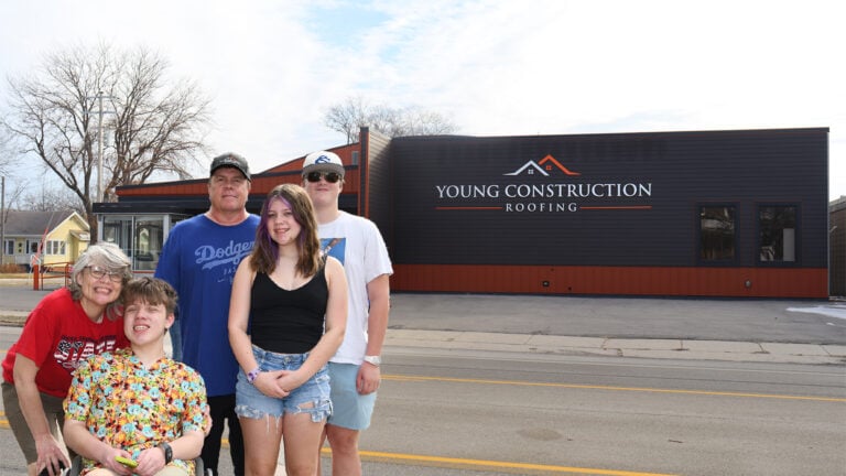 Five people smile outside Young Construction Roofing, standing before its branded building; trees and homes are in the background.
