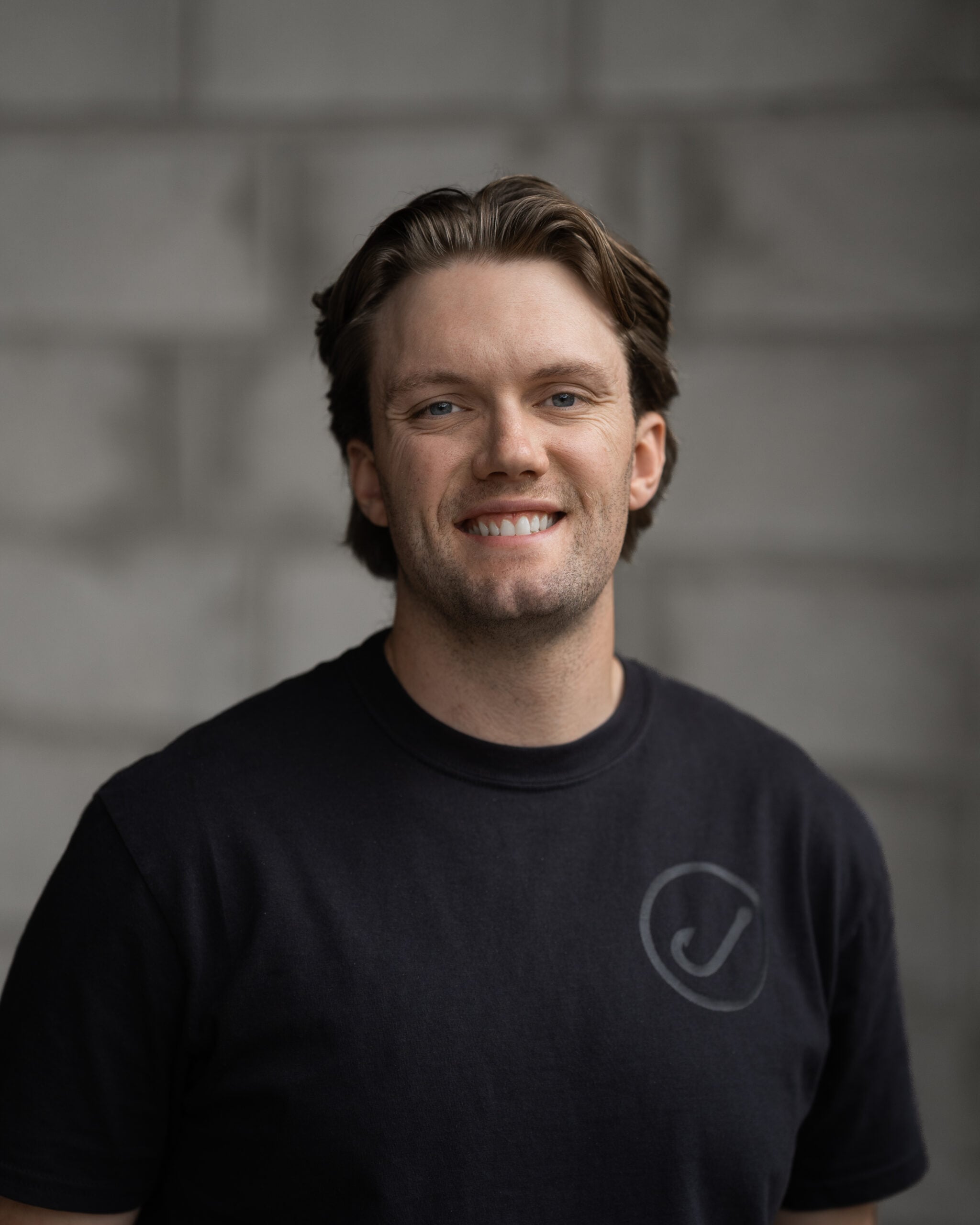 Zach Pattison, Hook Agency team member with short brown hair, smiles in a black logo tee against a blurred gray brick background.