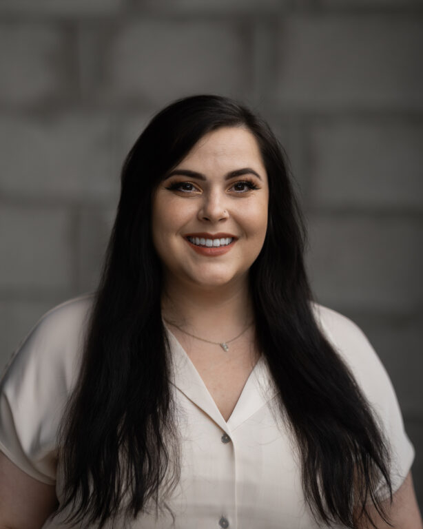 Justine Zaver, long dark hair and light blouse, stands smiling before a blurred gray brick wall for Hook Agency’s website.