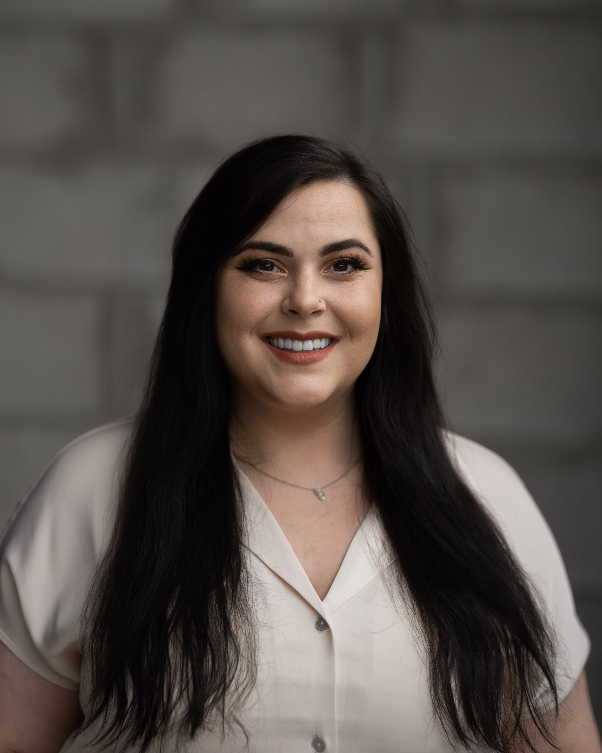 Justine Zaver, long dark hair and light blouse, stands smiling before a blurred gray brick wall for Hook Agency’s website.