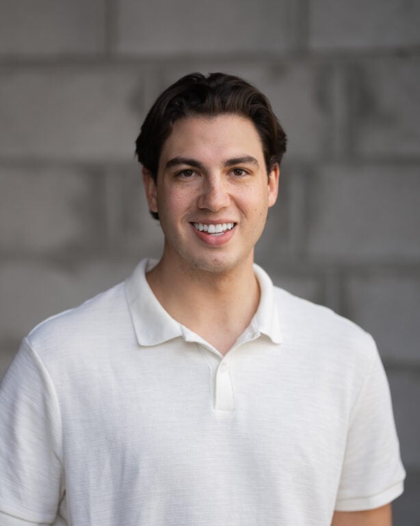 Jack Schmitz, team member at Hook Agency, stands smiling in a white collared shirt before a gray brick wall.
