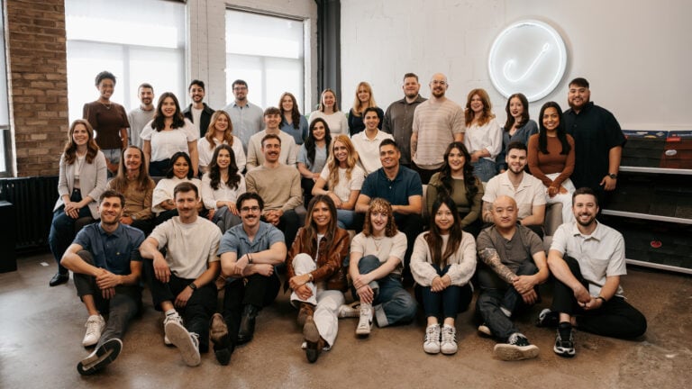 Meet Hook Agency’s team: 34 diverse professionals posing and smiling in a bright, modern office with a neon sign behind them.