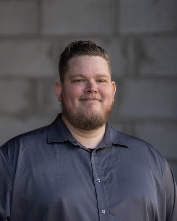 Vincent Baron, with short hair and a beard in a dark gray shirt, smiles confidently before a textured gray concrete wall.