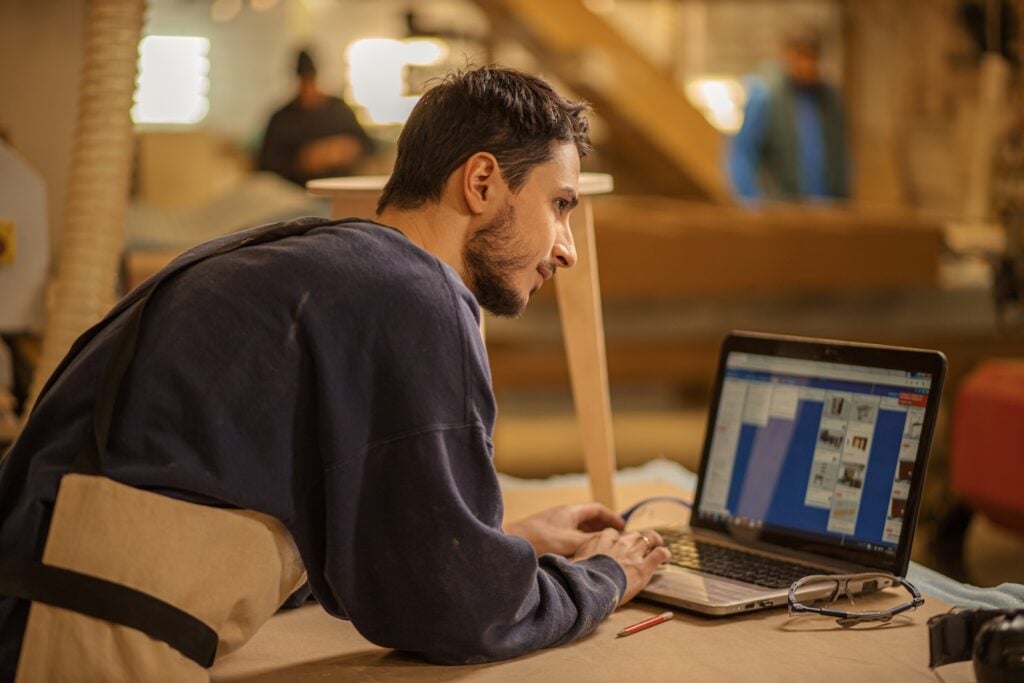 A contractor reviews top CRMs for home service businesses on a laptop at his workbench, tools nearby, others working behind him.