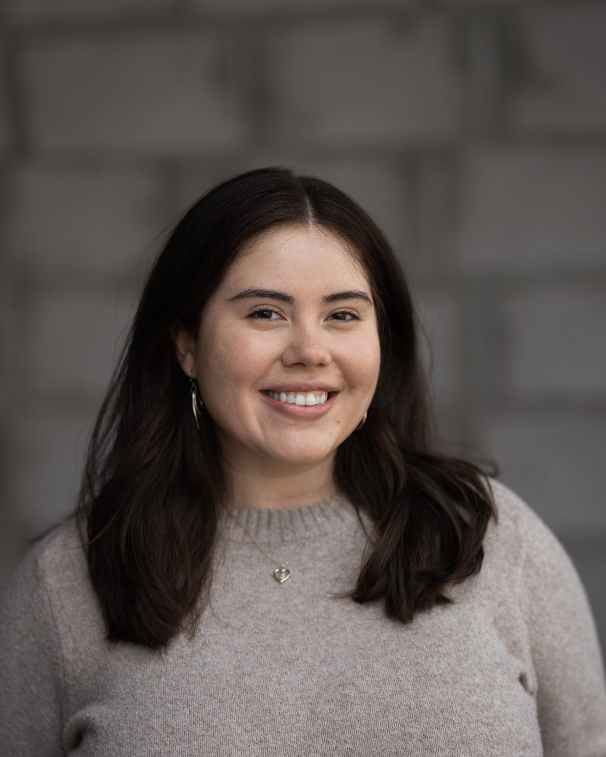 Andrea Obieta, a team member at Hook Agency with long dark hair, wearing a beige sweater and necklace, smiles confidently at the camera against a blurred gray brick wall background.