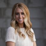 Smiling woman with long, wavy blonde hair in a white knit top stands before a blurred gray brick wall background.