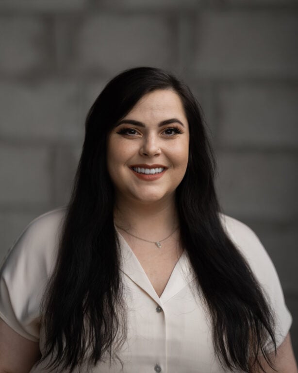 Justine Zaver, long dark hair and light blouse, stands smiling before a blurred gray brick wall for Hook Agency’s website.