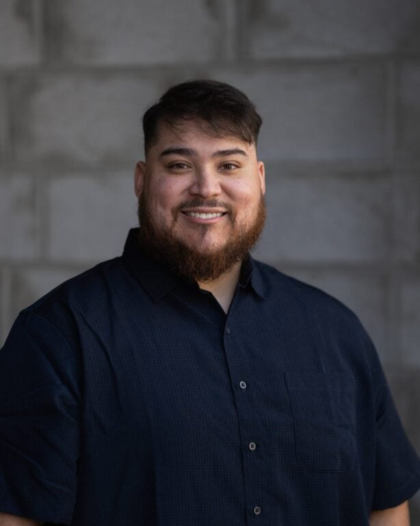 Joel Enrique Palomino, with short dark hair and beard, in a dark button-up shirt, smiling before a gray brick wall at Hook Agency.
