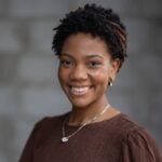 Mandelyn Miller of Hook Agency, with short curly hair and brown headband, smiles in a textured brown top and gold hoop earrings.