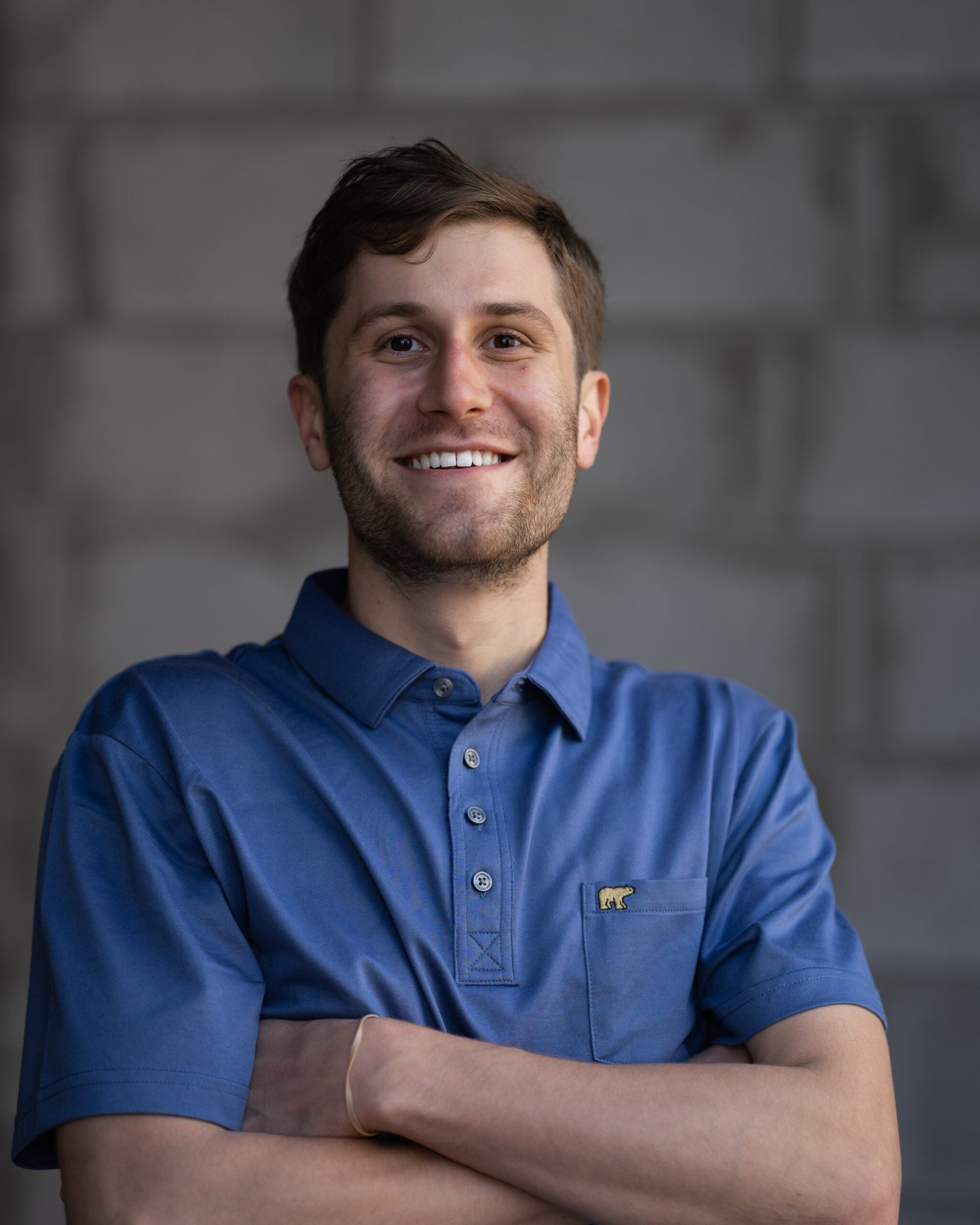 Confident young man with short brown hair in a blue collared shirt stands arms crossed, smiling, in front of a blurred brick wall.
