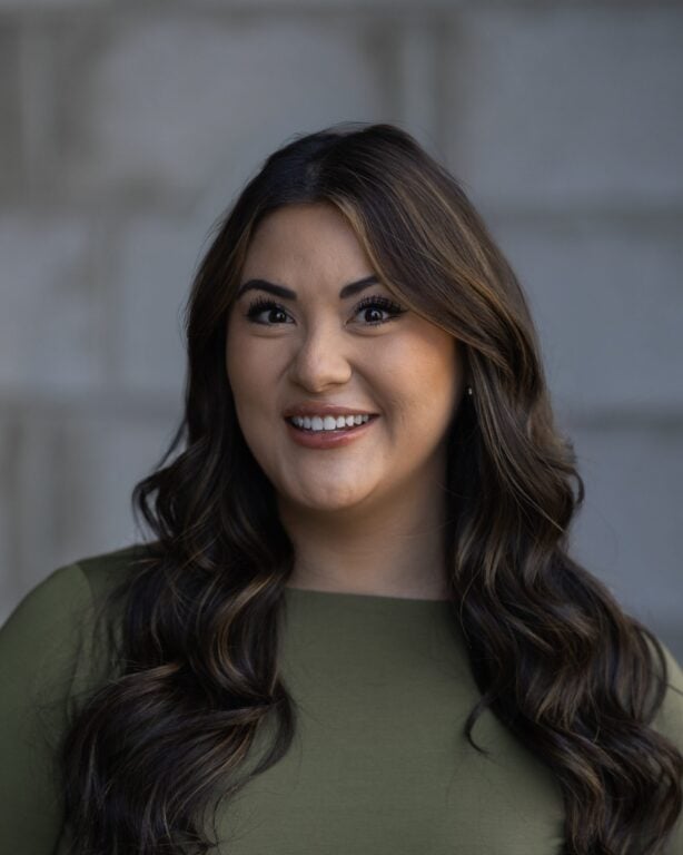 Sydnee Olsen, wearing a green top and with long wavy brown hair, smiles in front of a blurred gray background on Hook Agency’s site.