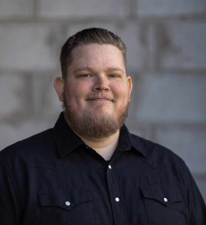 Vincent Baron, team member at Hook Agency, is pictured smiling in front of a blurred gray brick wall. He has a short beard, short hair, and wears a black button-up shirt.