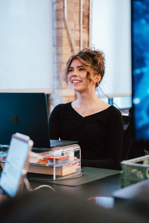 A professional woman with brown hair in a loose bun, dressed in a black top, sits at a desk with a laptop and books in a bright modern office, confidently preparing for her next task.
