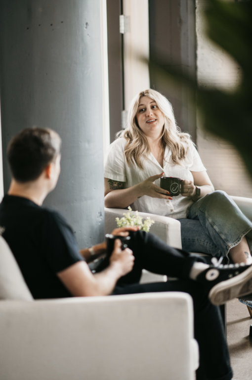 On Hook Agency’s website, two people sit in white chairs by a window, holding mugs and discussing career growth. The person closest to the camera wears a black shirt; the other has long blond hair and wears a white top with jeans. A small plant is placed on the table between them.