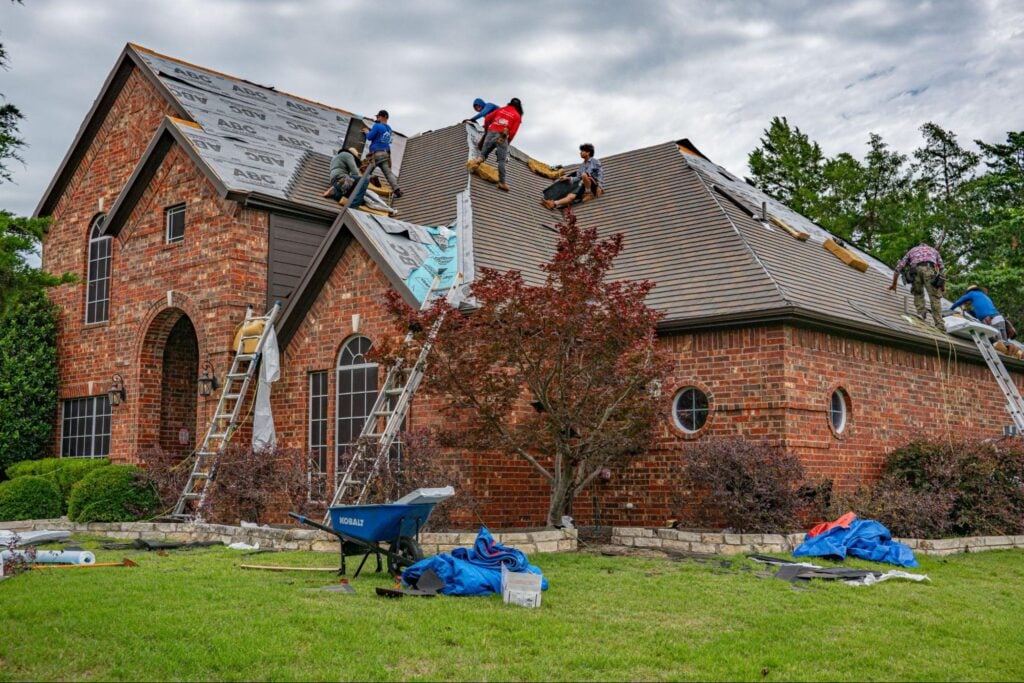 Professional roofing contractors install new shingles on a brick home, with tarps and tools organized on the lawn below.