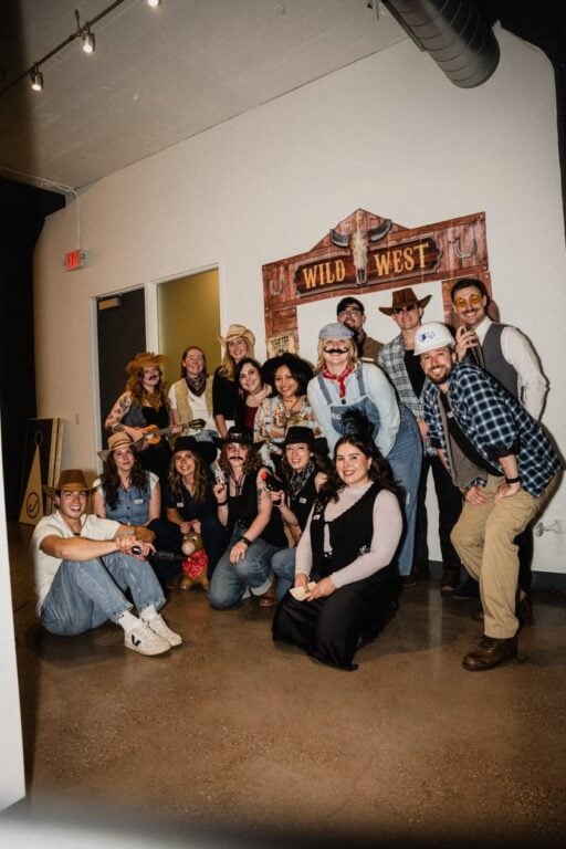 Hook Agency team members, dressed in cowboy and cowgirl costumes that reflect their unique roles and personalities, pose and smile together indoors before a "Wild West" sign.