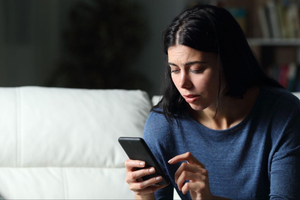 A woman with long dark hair sits on a white couch, wearing a blue top, indoors. She looks concerned while intently checking her smartphone for plumbing leads or urgent job notifications.