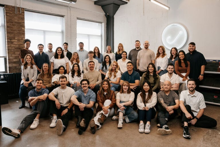 The Hook Agency team—32 members—gather in a modern office with a neon circular sign and large windows. Some are seated on the floor, others stand behind, all smiling at the camera.
