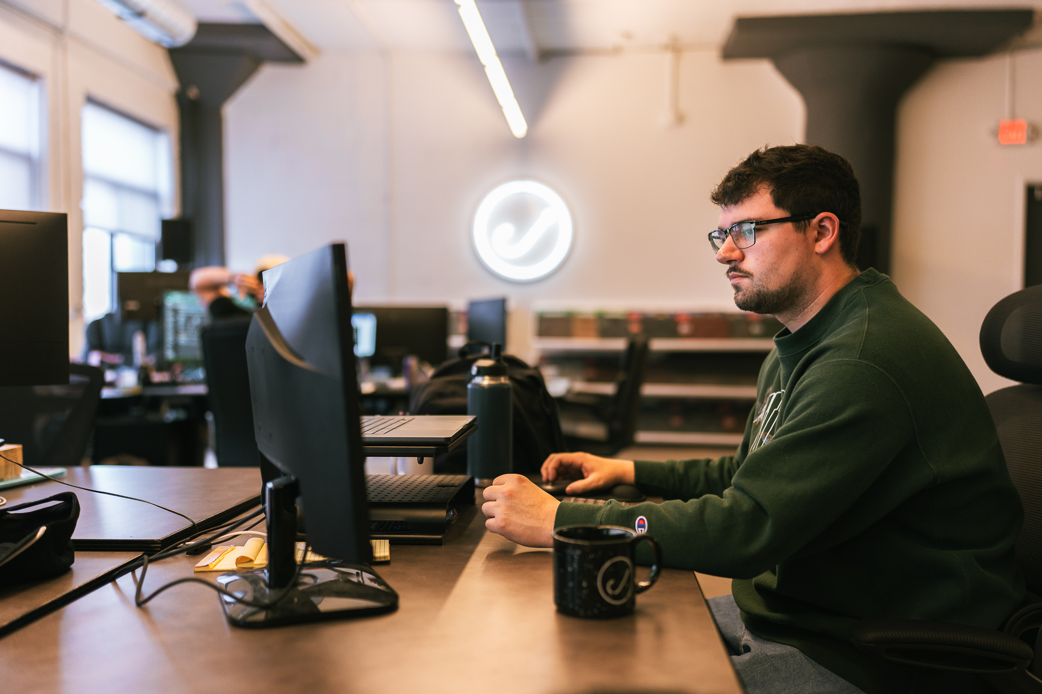 A man in glasses and a green sweatshirt works at his computer in Hook Agency’s modern office, surrounded by desks, monitors, and a coffee mug. A circular neon sign on the back wall highlights the agency’s focus on inspiring professional growth.