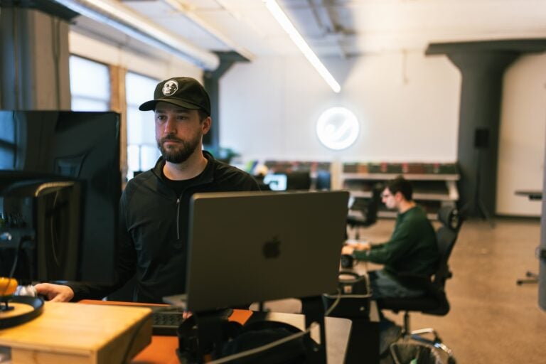 In Hook Agency’s modern, open office, a man in a black cap works at a standing desk with a laptop and monitors, while another team member focuses at a seated workstation in the background.