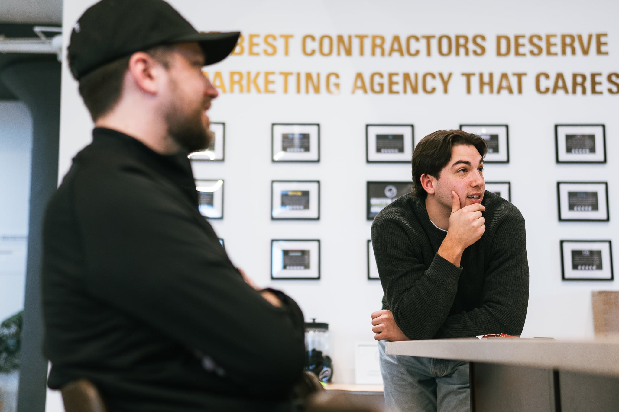 At Hook Agency’s office, two men discuss a project; one listens attentively while leaning on a counter, and the other appears blurred in the foreground. Motivational quotes and framed photos are visible on the back wall.