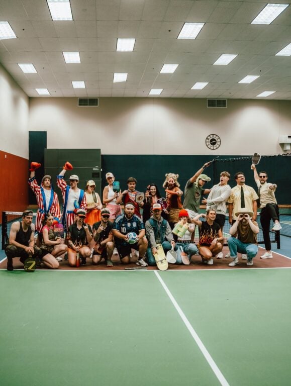 Team members in vibrant, unconventional costumes stand together on an indoor tennis court, smiling and making playful gestures, demonstrating Hook Agency’s creative culture and diverse talent.