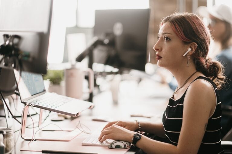 A professional with red hair, wearing wireless earbuds and a striped top, reviews paid ads management on her monitor at an office desk; colleagues work in the background, illustrating Hook Agency’s digital marketing expertise.