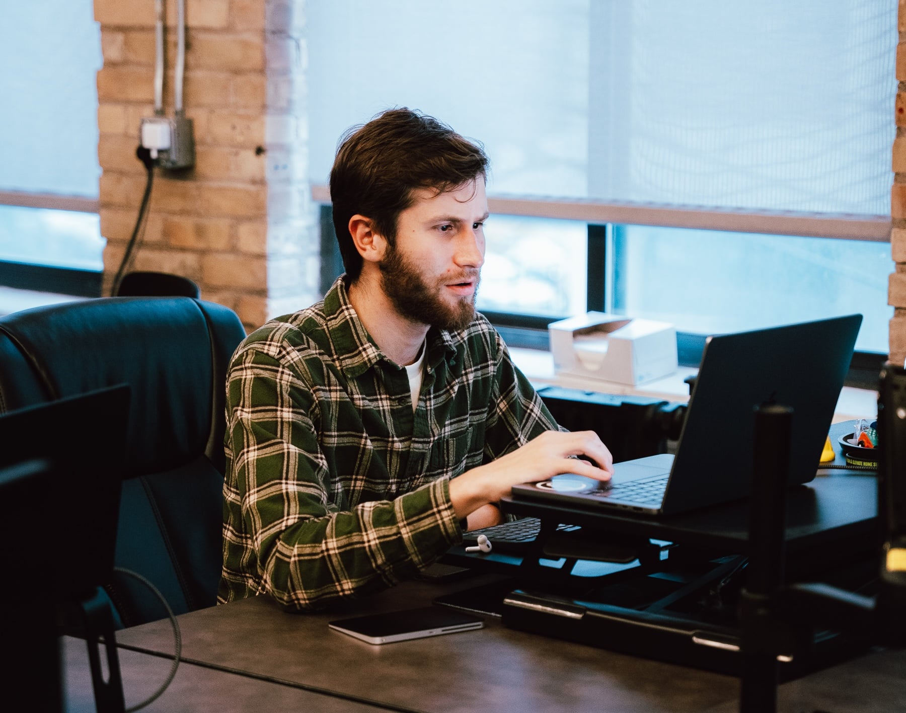 A bearded man in a green plaid shirt sits at a desk in a modern office with brick walls and large windows, working on a laptop and focusing on PPC for Home Services and paid ads management for Hook Agency.