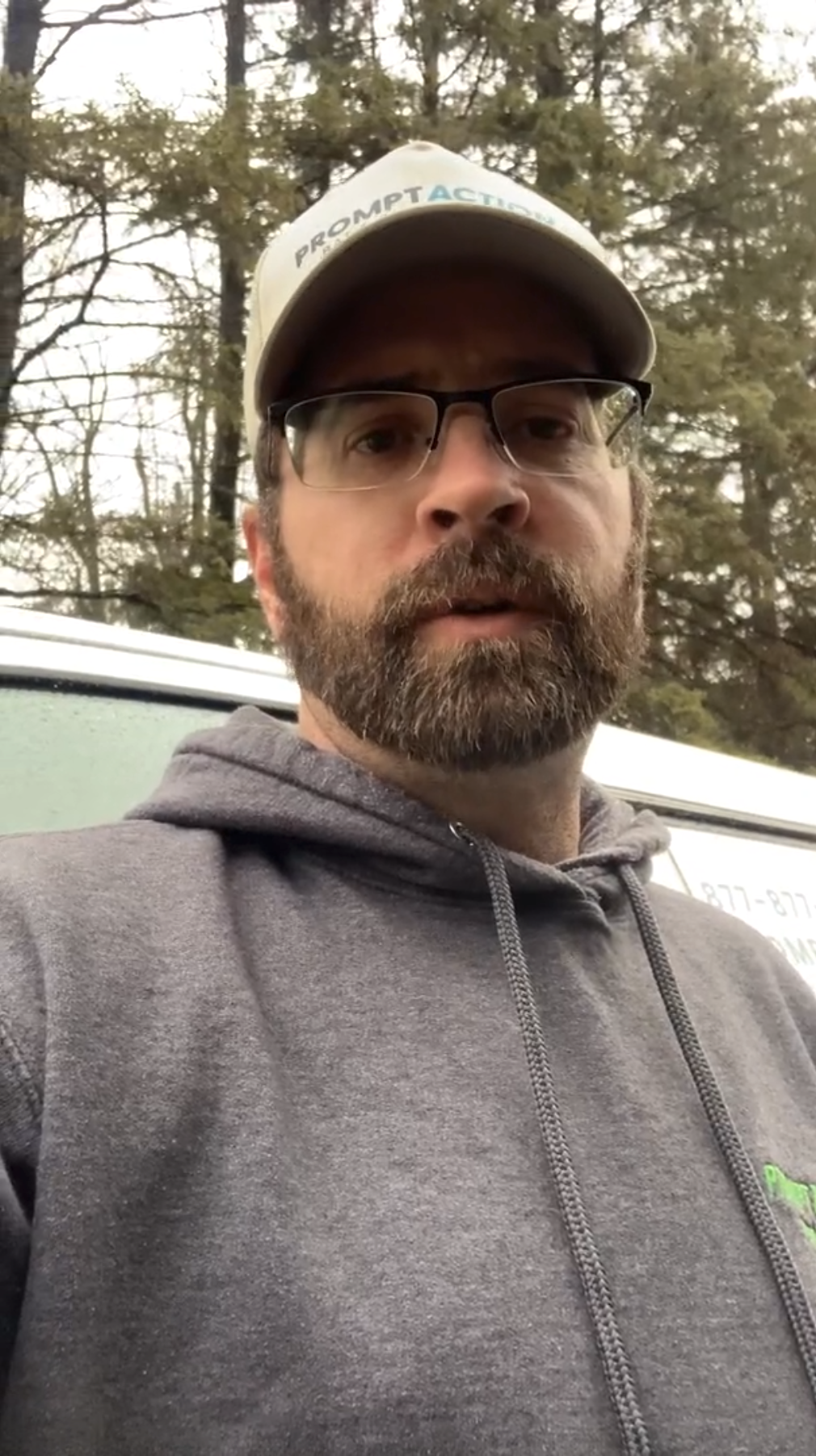 A bearded man in glasses, a cap, and a gray hoodie stands outside in front of a white vehicle and trees, representing Hook Agency’s approachable team.