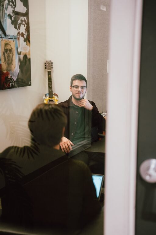 A Hook Agency SEO consultant sits across from a client in a creative workspace, featuring a guitar and vibrant artwork on the wall. Viewed through a glass door, the scene highlights an active strategy session focused on results-driven SEO solutions.