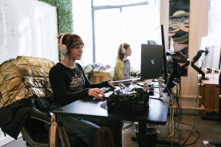 Hook Agency team members work at computers in a sunlit, modern office. The person in the foreground, wearing headphones, concentrates on building custom websites for home service businesses; another team member sits further back.