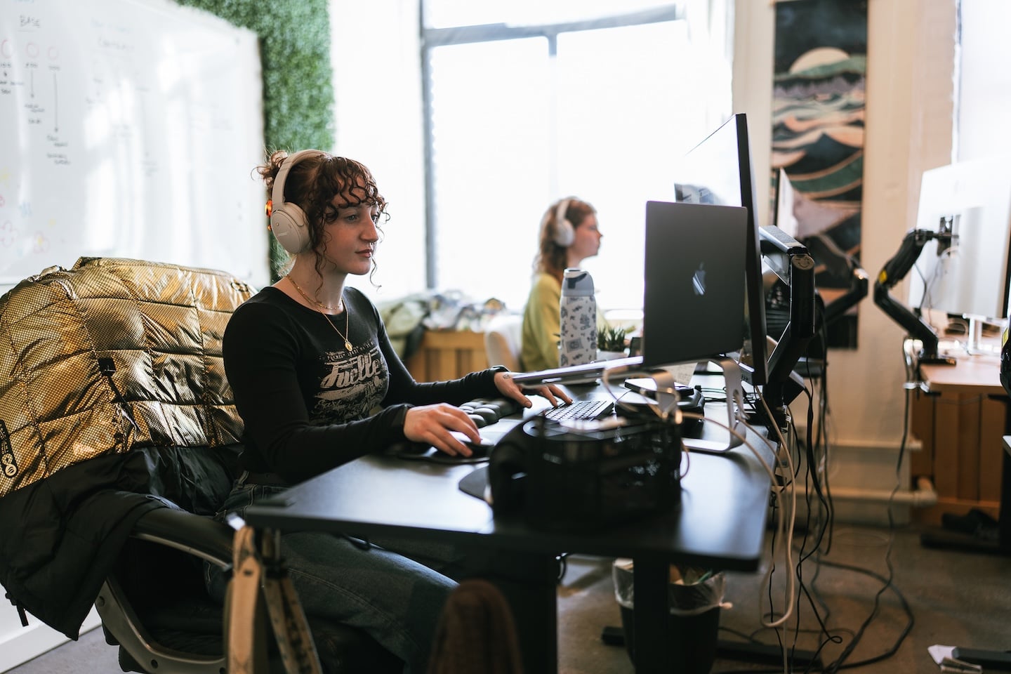 Hook Agency team members work at computers in a sunlit, modern office. The person in the foreground, wearing headphones, concentrates on building custom websites for home service businesses; another team member sits further back.