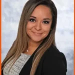 Angelica Brager, team member at Hook Agency, is pictured with long straight hair, wearing a black blazer over a light top, smiling confidently in a professional studio portrait against a light gray background.