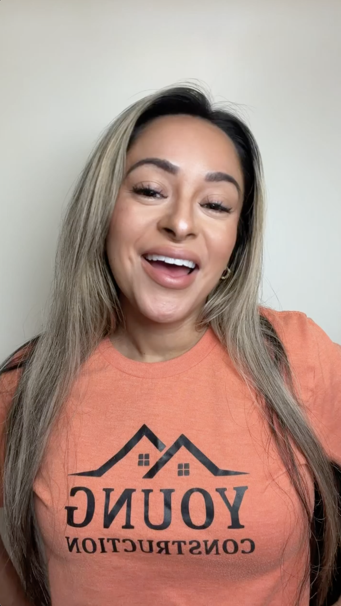 A smiling woman with long blonde hair wears an orange "Young Construction" t-shirt and stands against a light-colored wall, representing Young Construction on Hook Agency's website.